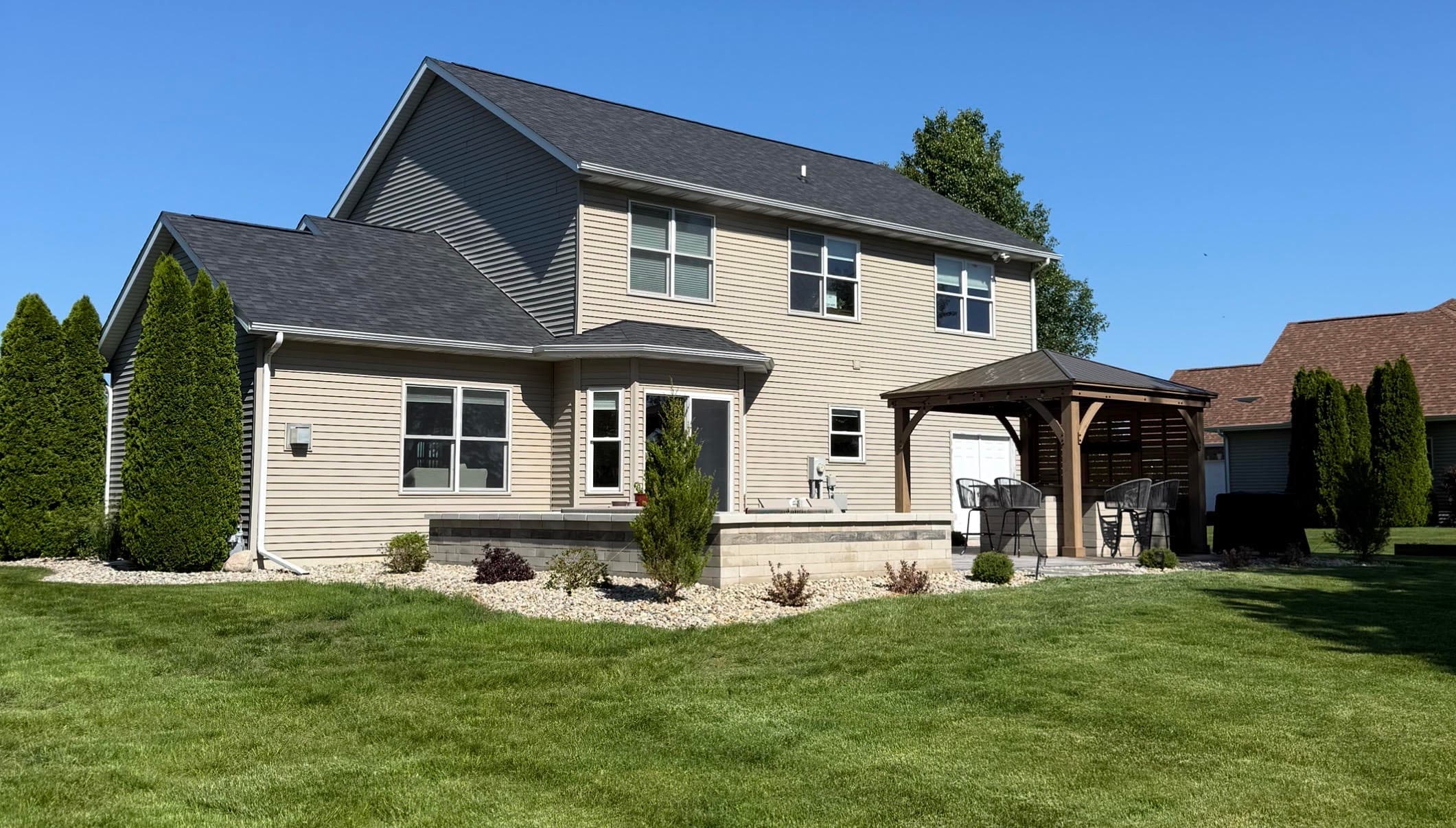 Two-story beige house with a patio, gazebo, and landscaped yard on a sunny day.