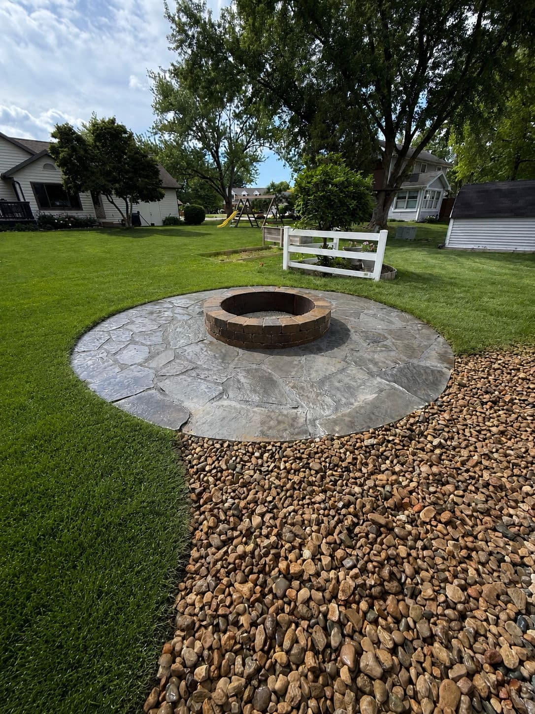 Fire pit surrounded by stone patio and gravel in a well-maintained backyard.