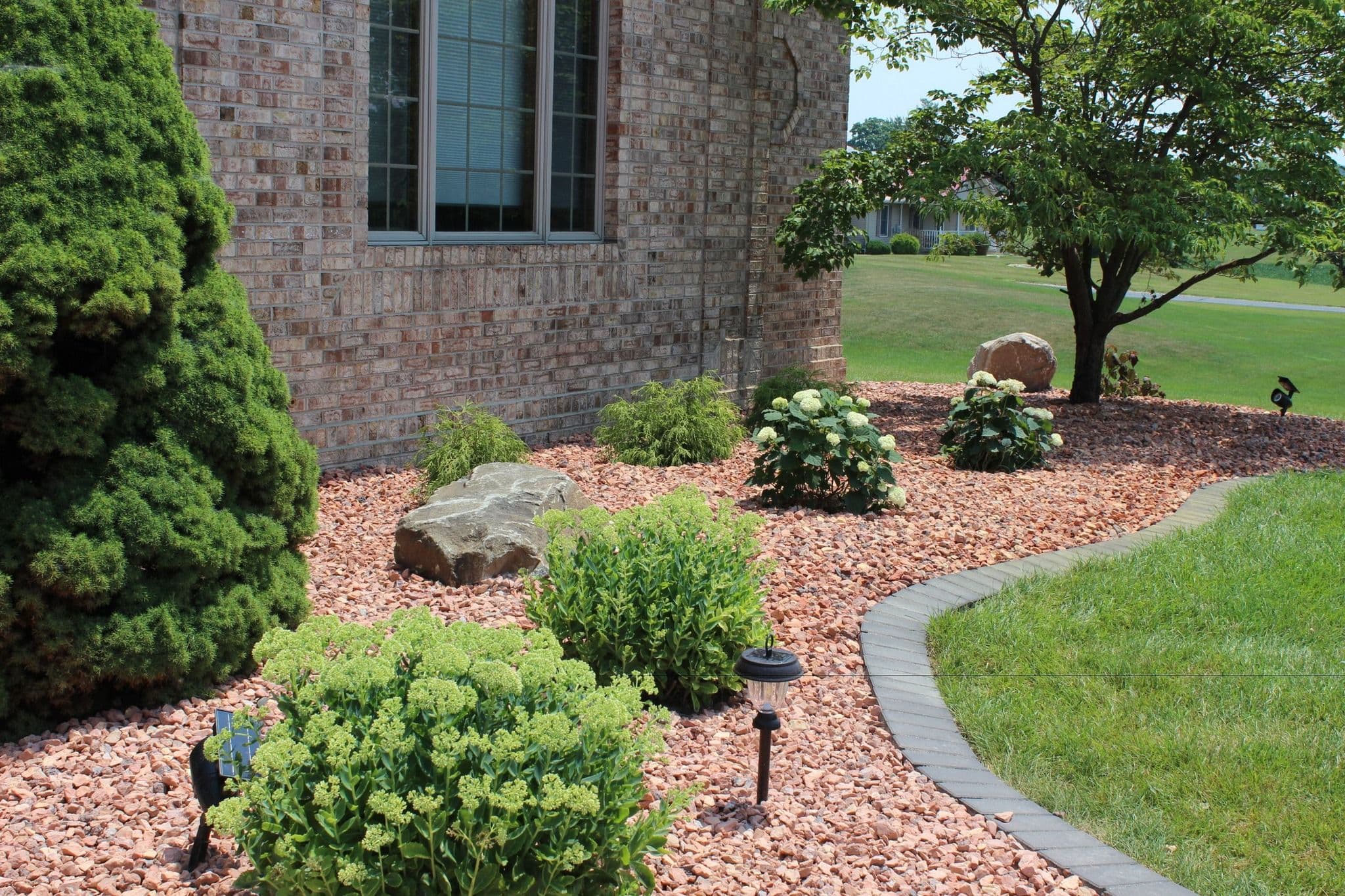 Lush garden with stone path, shrubs, and decorative rocks beside a brick home.