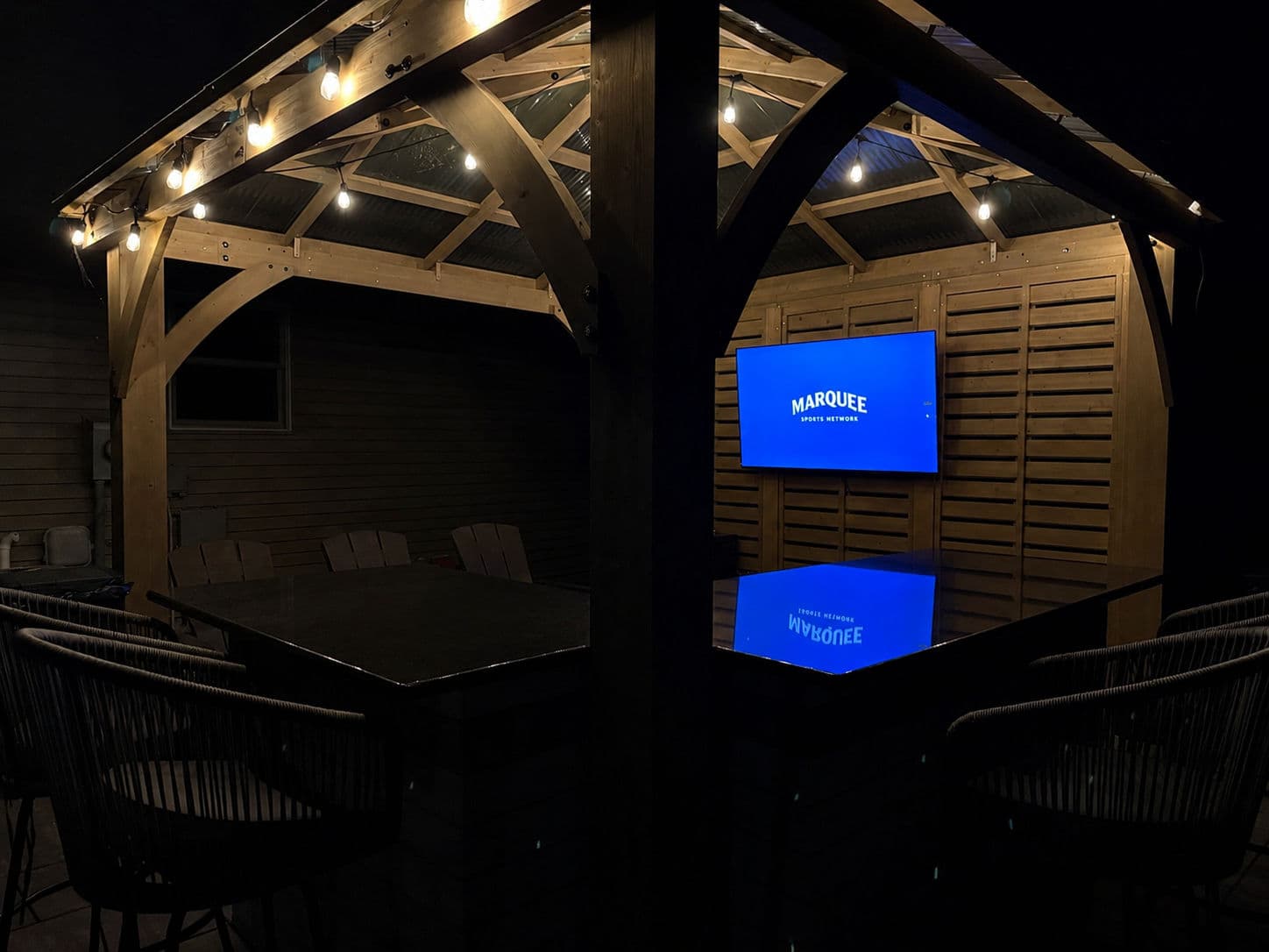 Wooden gazebo with string lights and a large screen displaying "MARQUEE" at night.