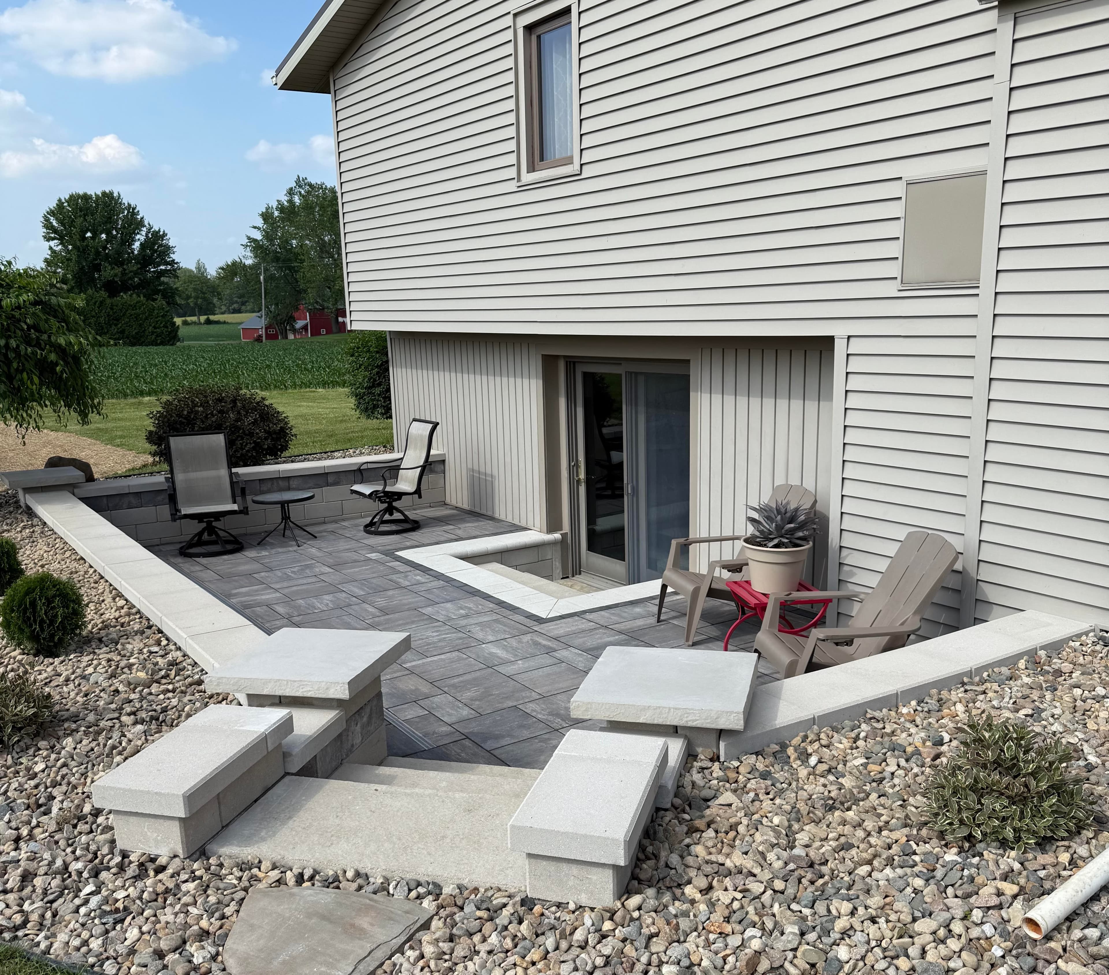 Modern outdoor patio with seating, stone pavers, and landscaped greenery beside a house.