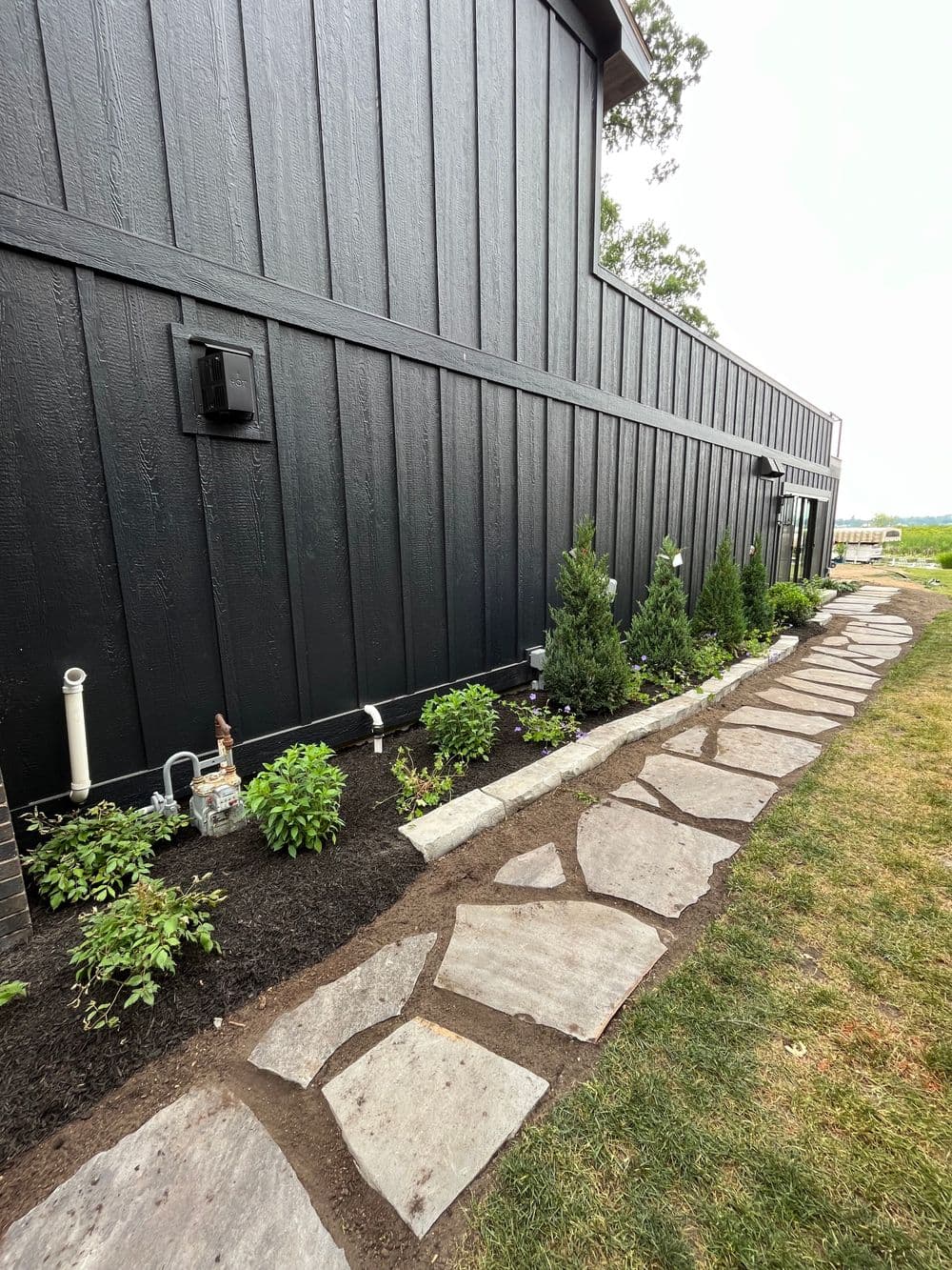 Stone pathway alongside a black wooden wall, featuring landscaping with shrubs and small trees.