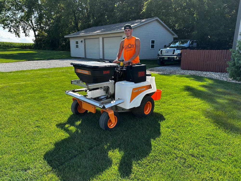 Person operating a spreader on a lawn, with a garage and truck in the background.