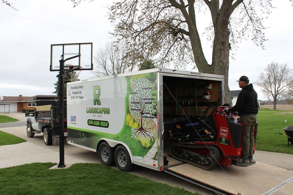 Landscaping trailer with equipment and worker in residential yard. Tree care services displayed.