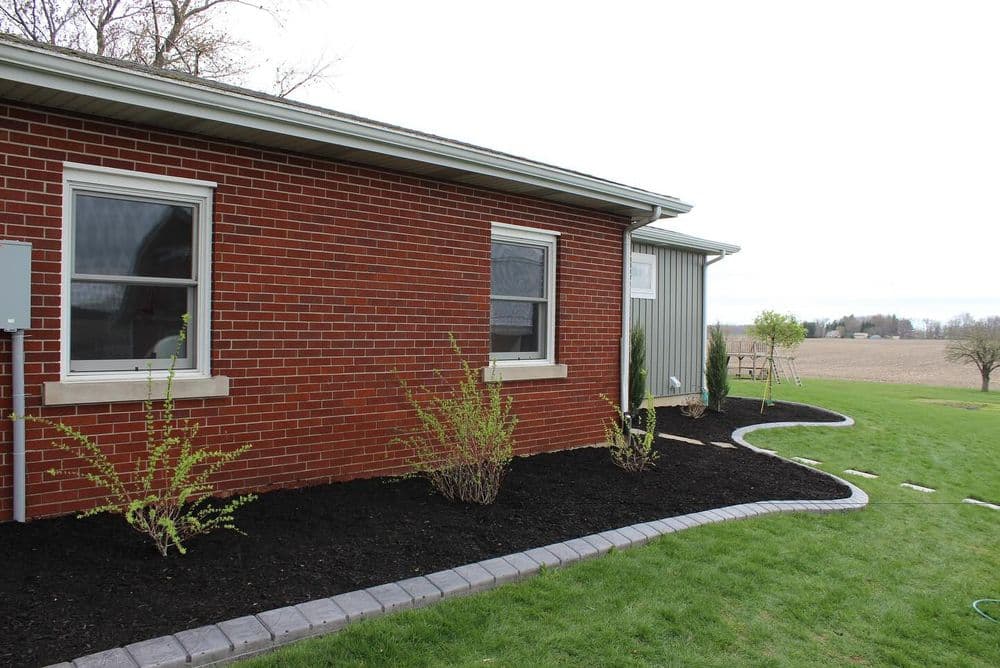 Brick house exterior with landscaped garden, mulch beds, and open field background.