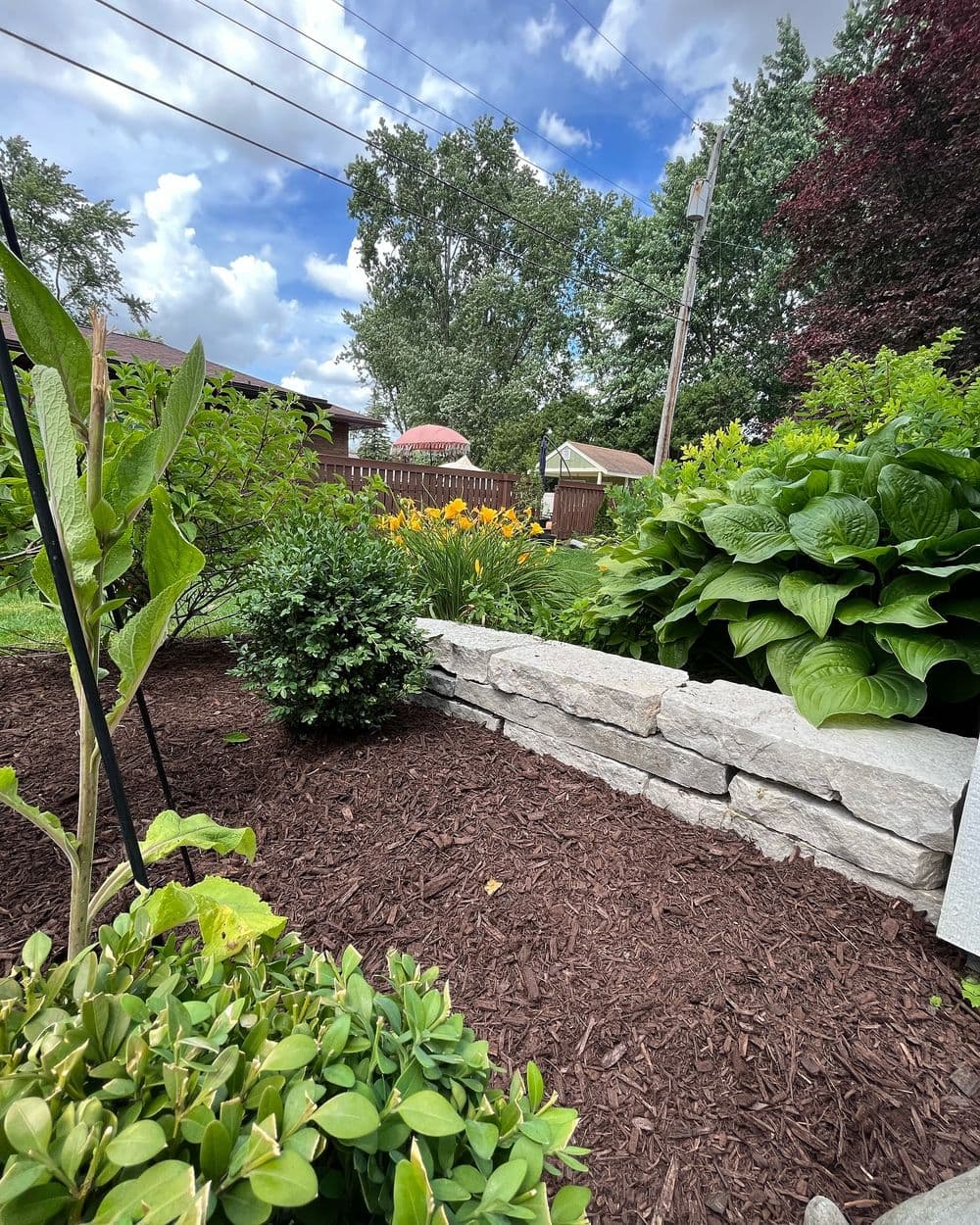 Lush garden with stone wall, vibrant flowers, greenery, and blue sky in the background.