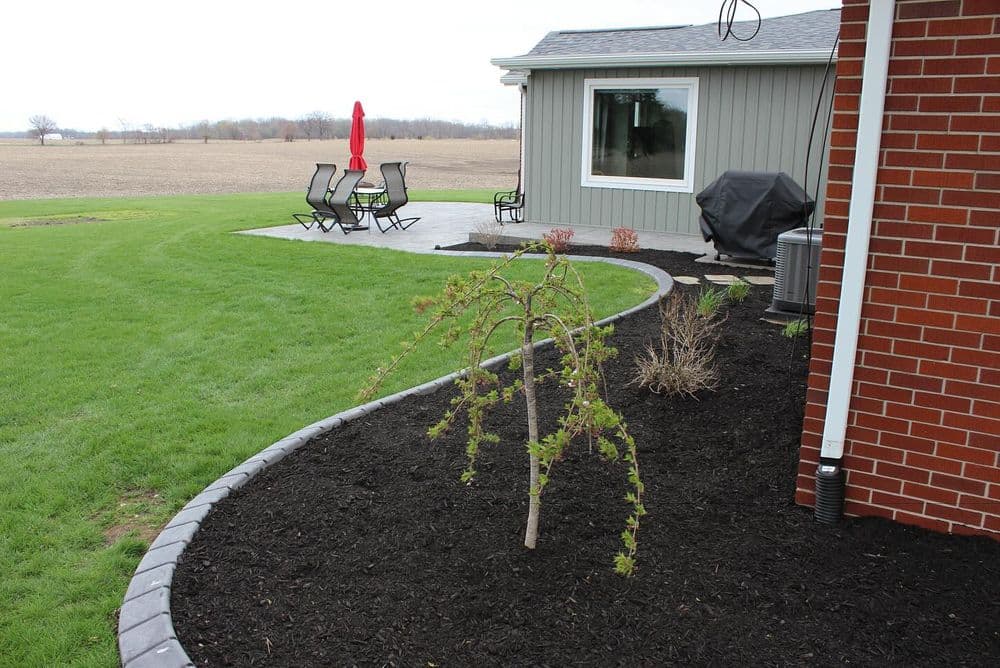 Newly landscaped yard with small tree, patio furniture, and a grill beside a brick house.